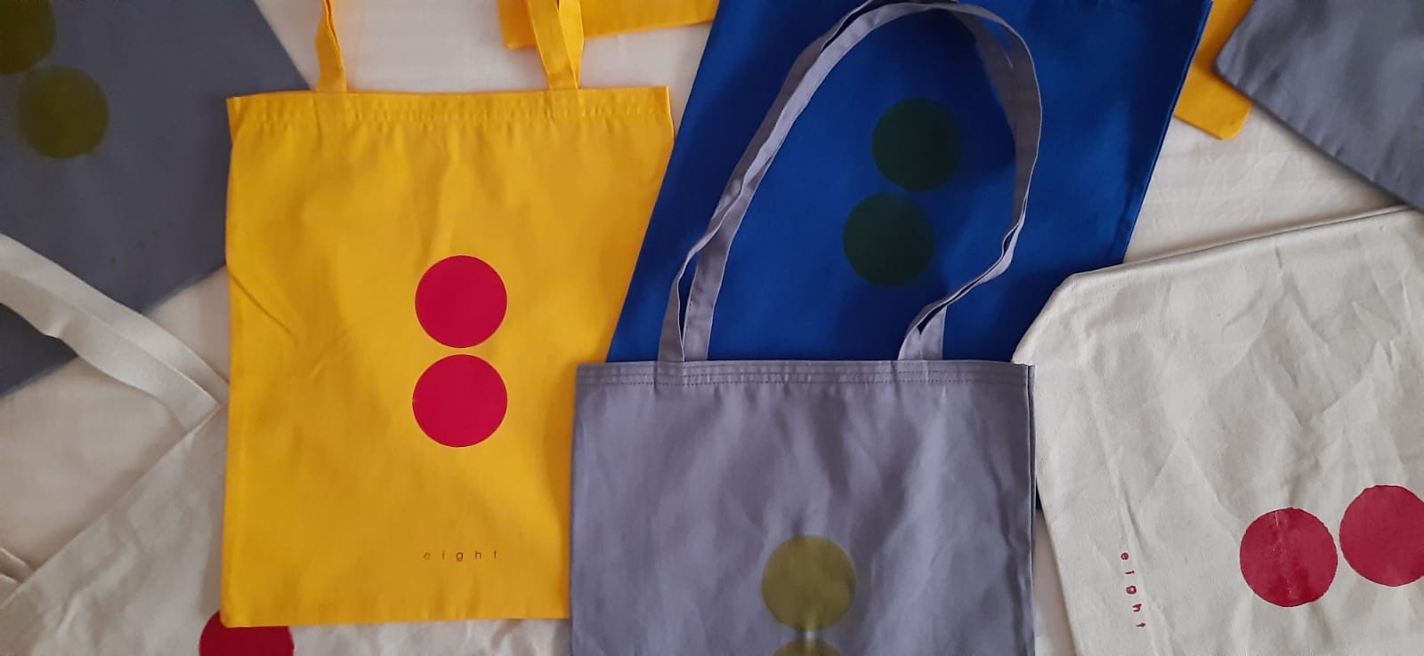 Collections-Lets-Celebrate-Eight-Hamide-Design-Studio Eight totebags of a variety of colors are spread around overlapping one another.